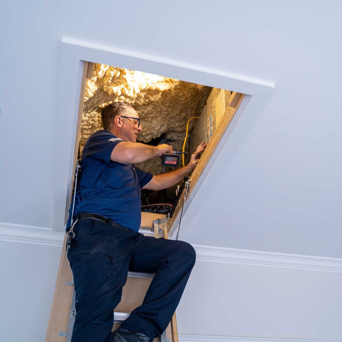 A man wearing glasses and a blue shirt works in an attic space, accessed via a pull-down ladder. He is using tools and appears to be installing or inspecting wiring near insulation.